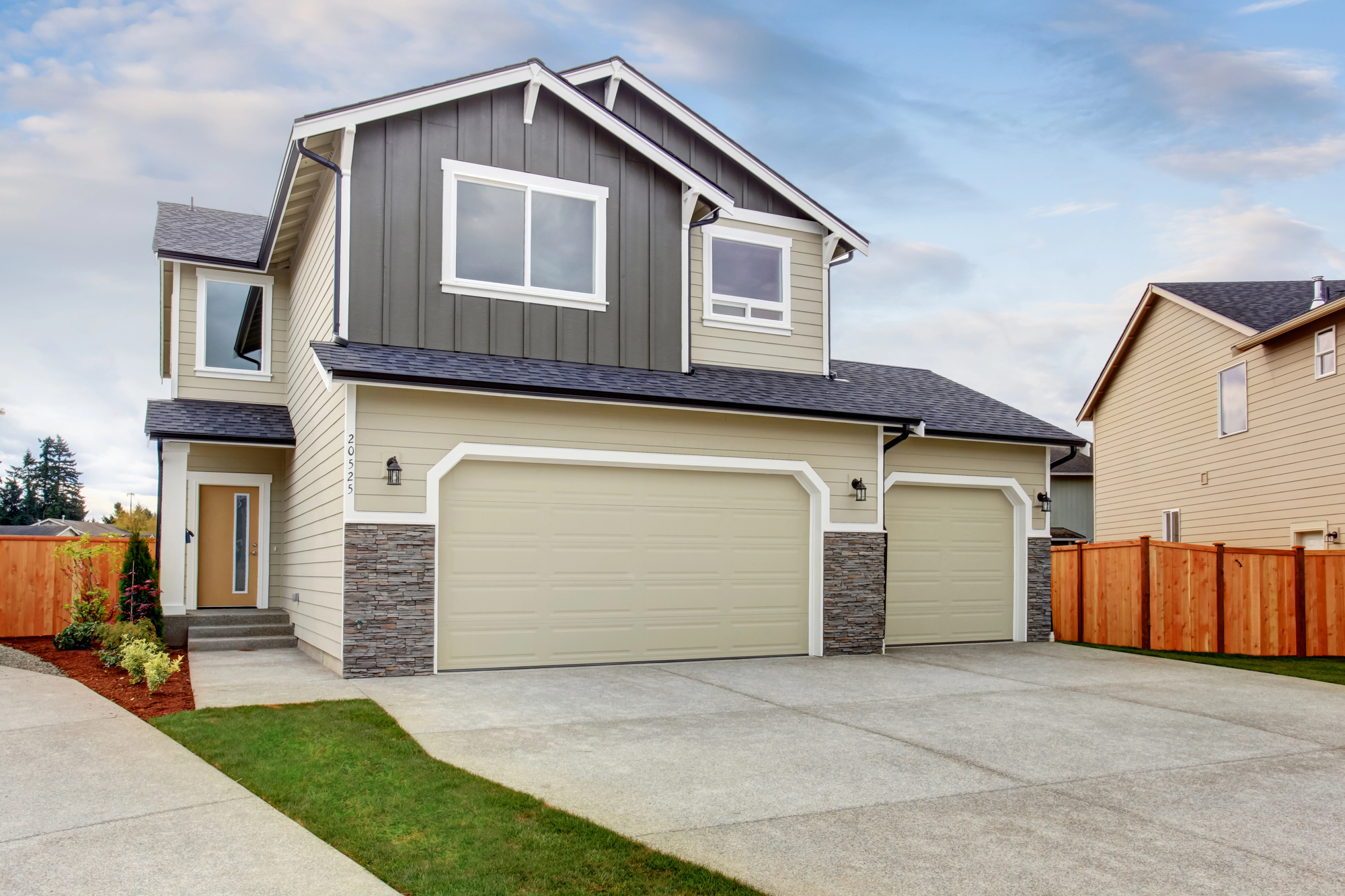 Two-story suburban house with gray siding, stone accents, and three-car garage, set on a concrete driveway with a wooden fence and well-kept lawn.