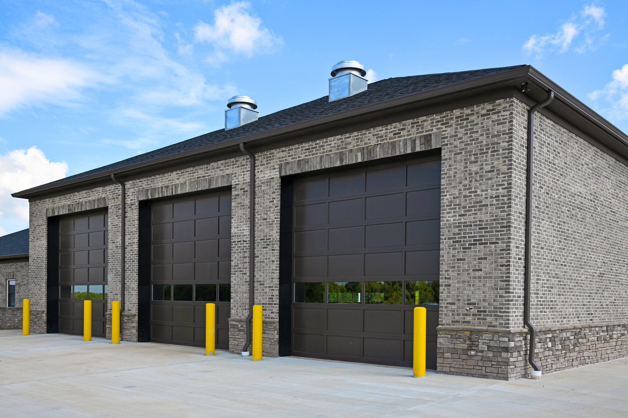 A modern brick building with three large, closed garage doors and yellow safety bollards in front, under a blue sky with clouds.