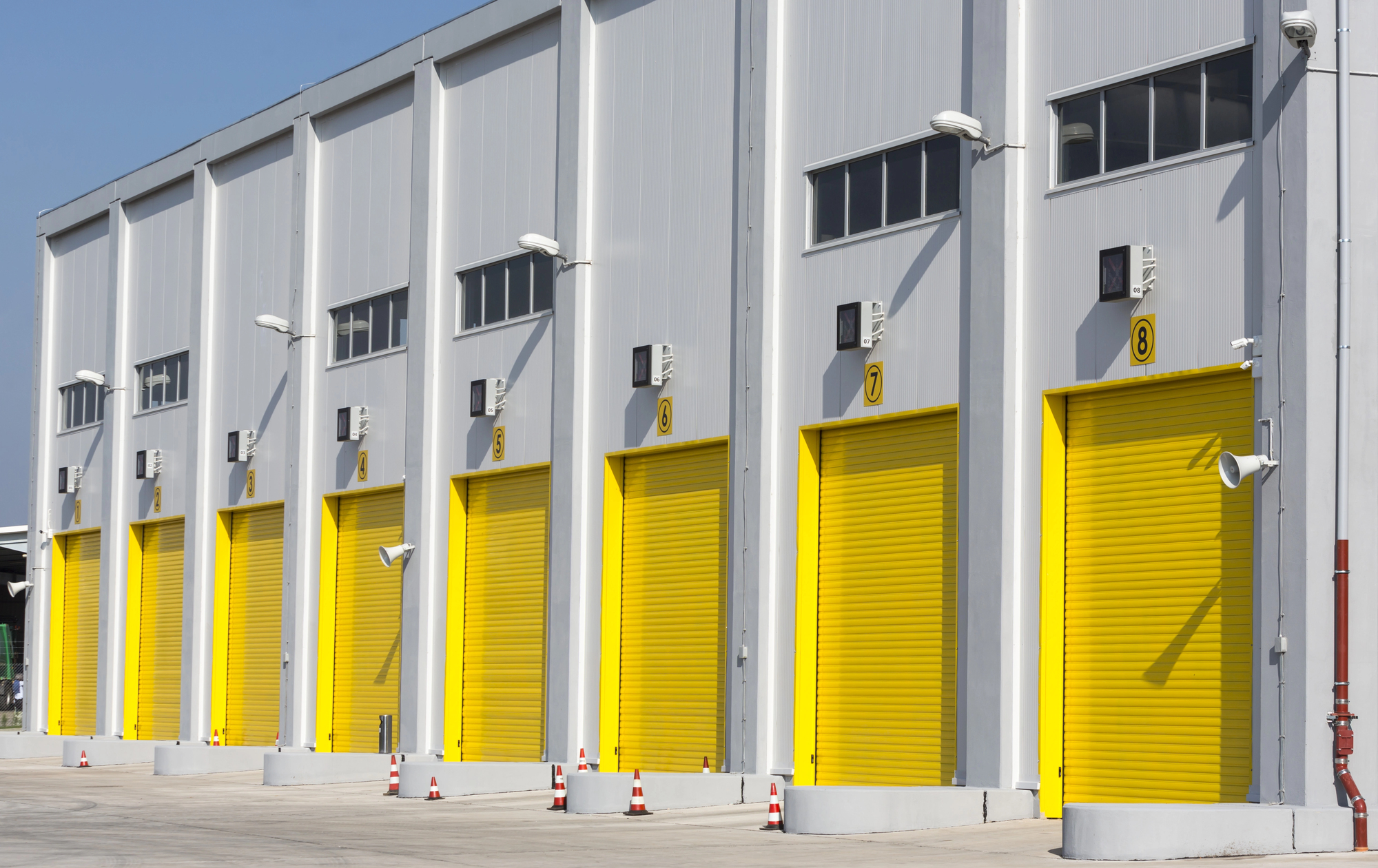 A row of industrial building loading dock doors, each painted bright yellow and numbered, with traffic cones placed in front.