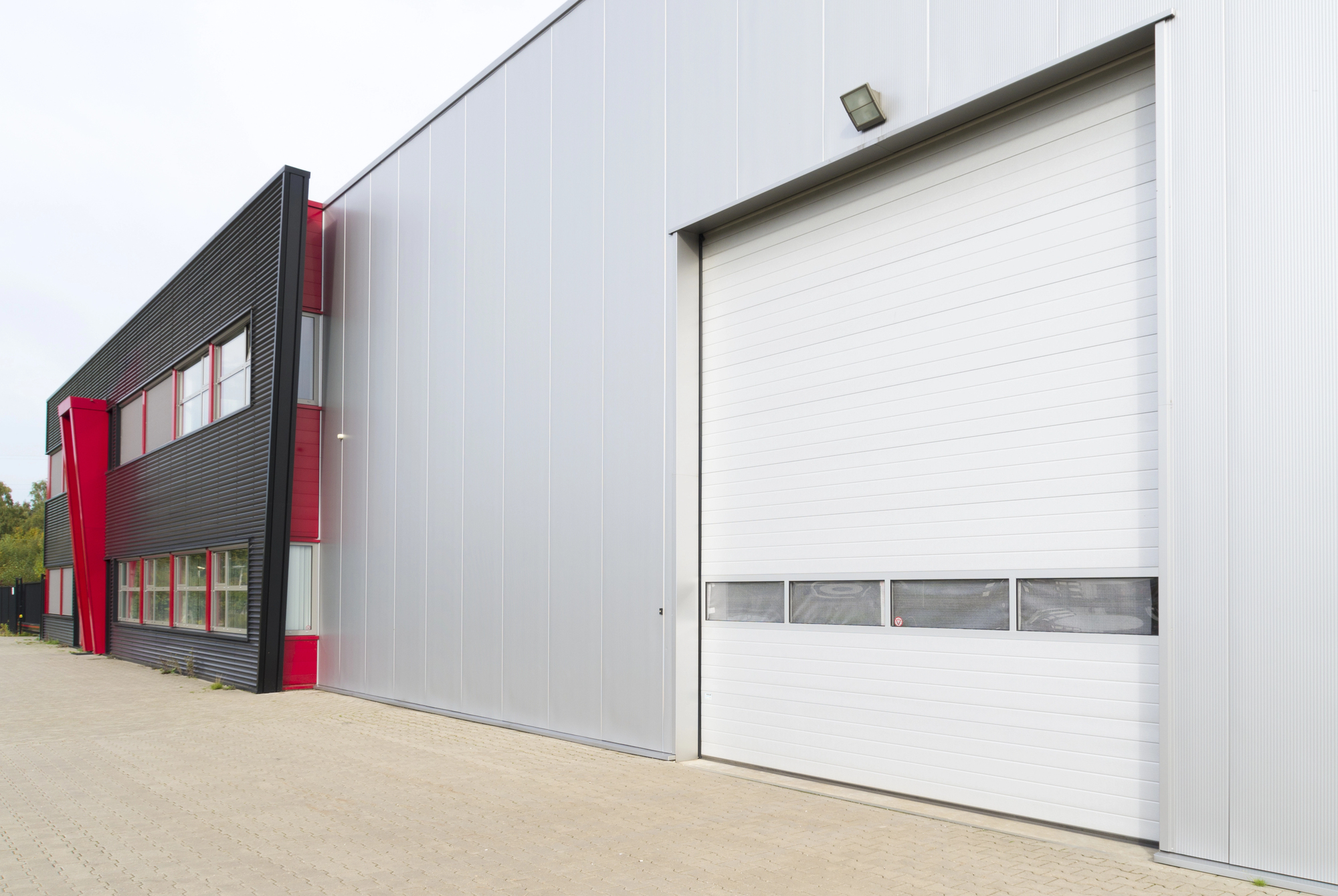 Modern industrial building exterior with large white roll-up garage door, red and black accents, and paved outdoor area.