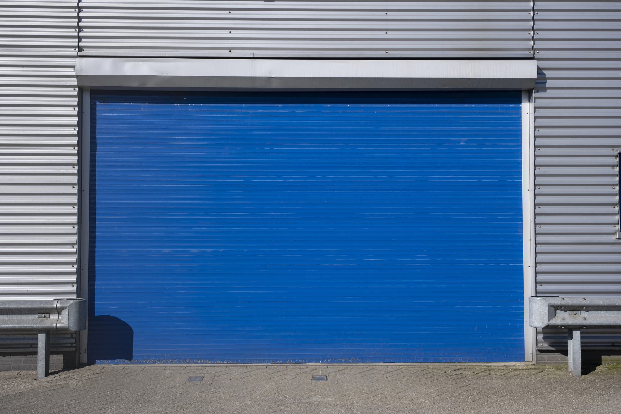 A closed blue industrial garage door set in a corrugated metal building, with pavement in front and metal guardrails on each side.