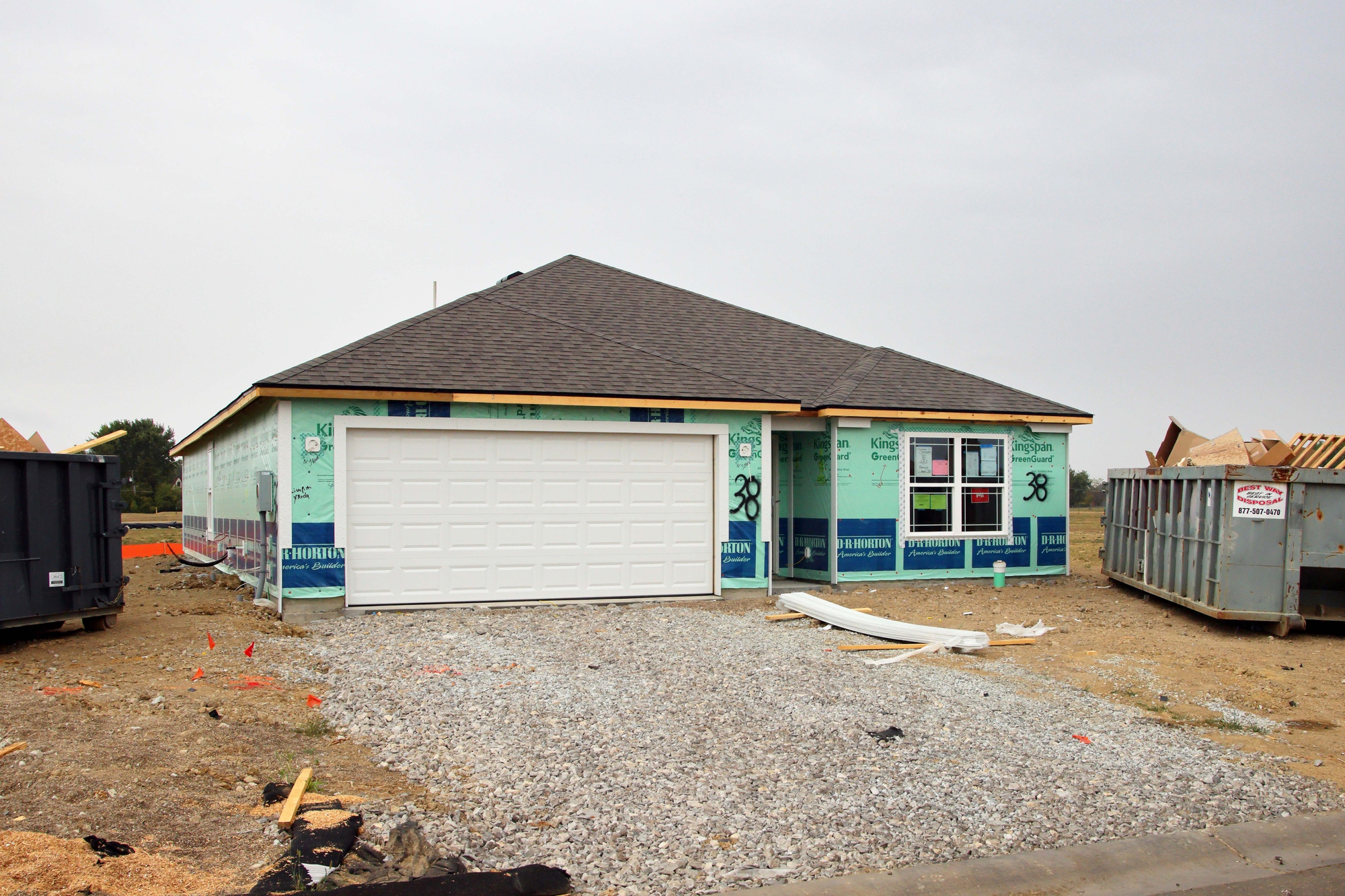 A single-story house under construction with exterior insulation visible, a white garage door, gravel driveway, and a dumpster nearby on a cloudy day.