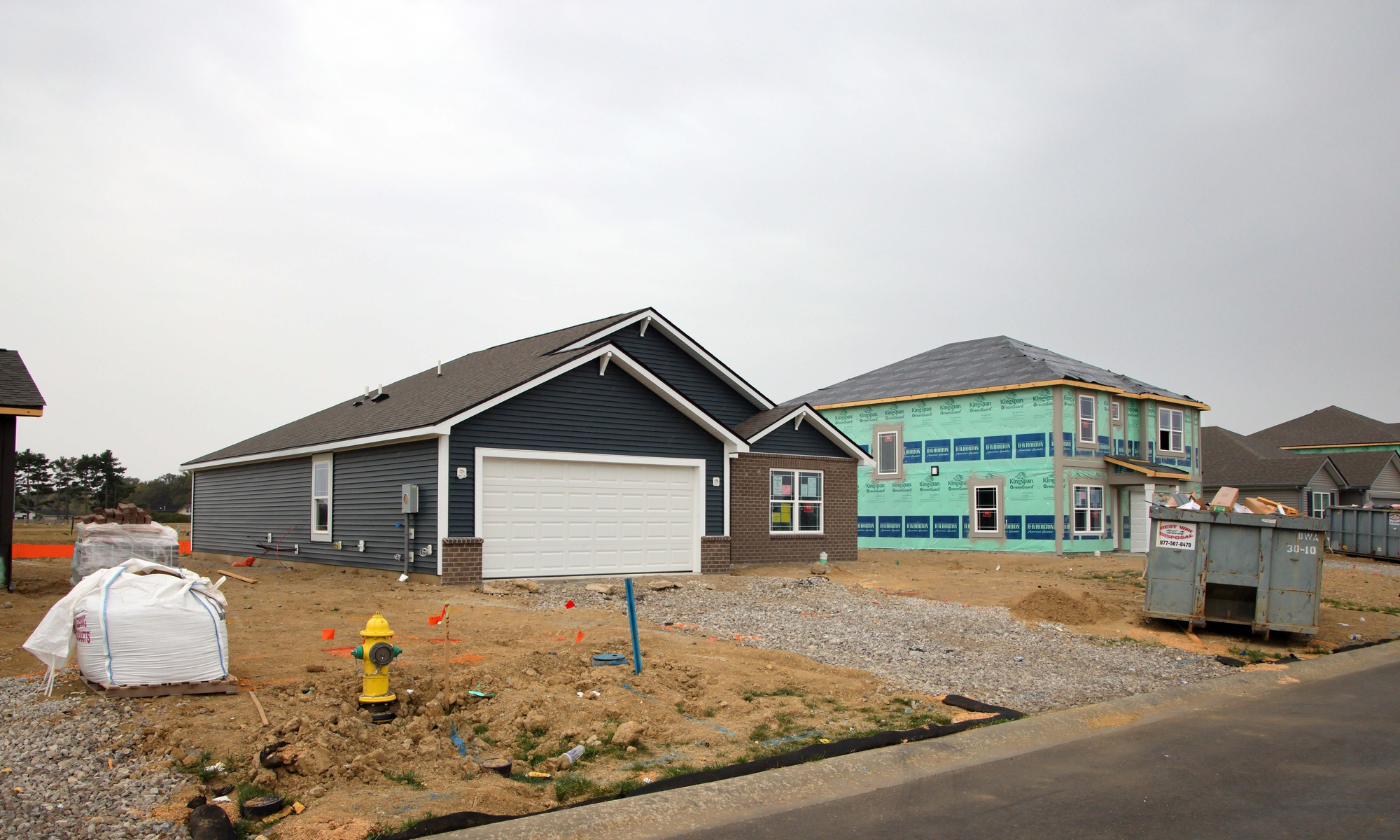 Two houses under construction in a suburban neighborhood, one mostly finished with dark siding, the other wrapped in green insulation material, with building materials and a dumpster nearby.