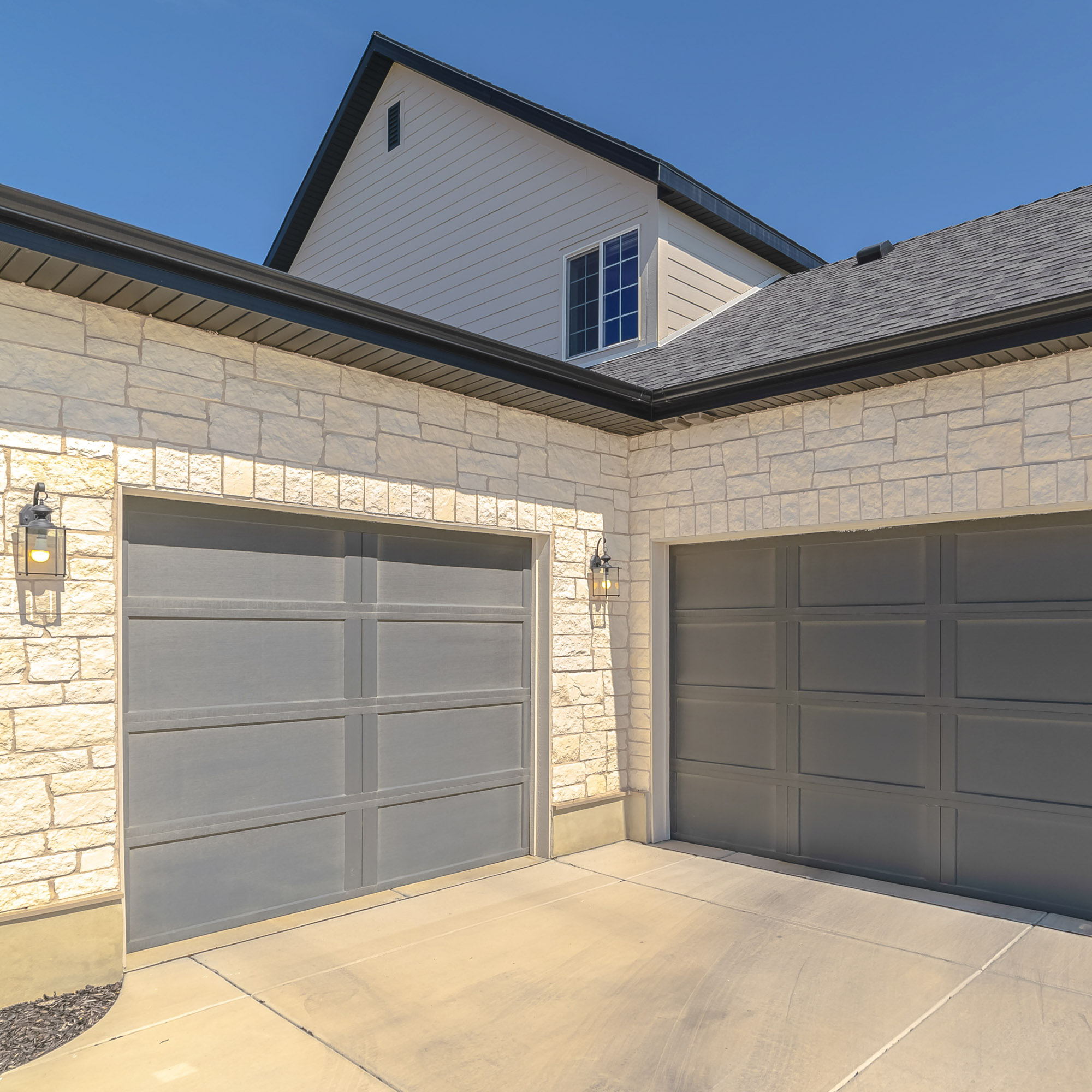 Two closed gray garage doors on a modern house with light stone exterior under a clear blue sky.