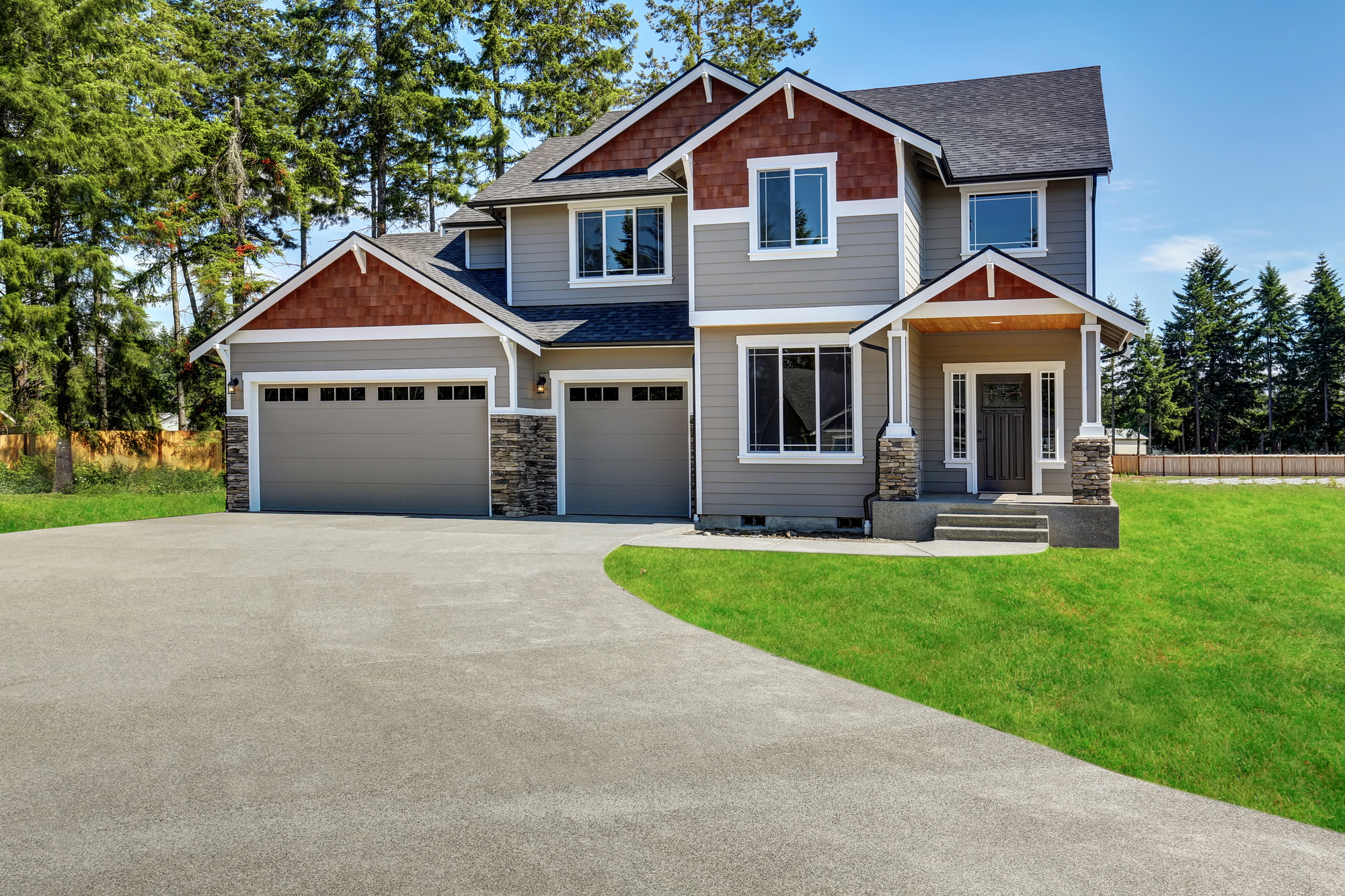 Two-story modern suburban house with gray siding, stone accents, a three-car garage, and a large driveway surrounded by green lawn and trees.