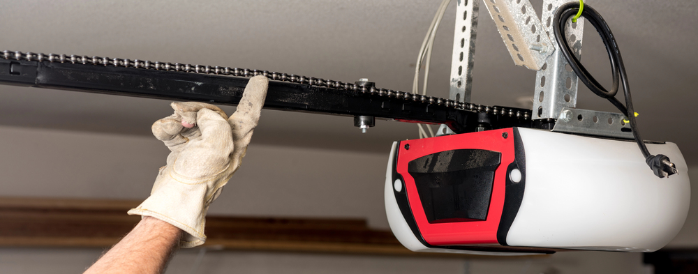 A person wearing a glove adjusts or inspects the chain mechanism of an automatic garage door opener mounted on the ceiling.