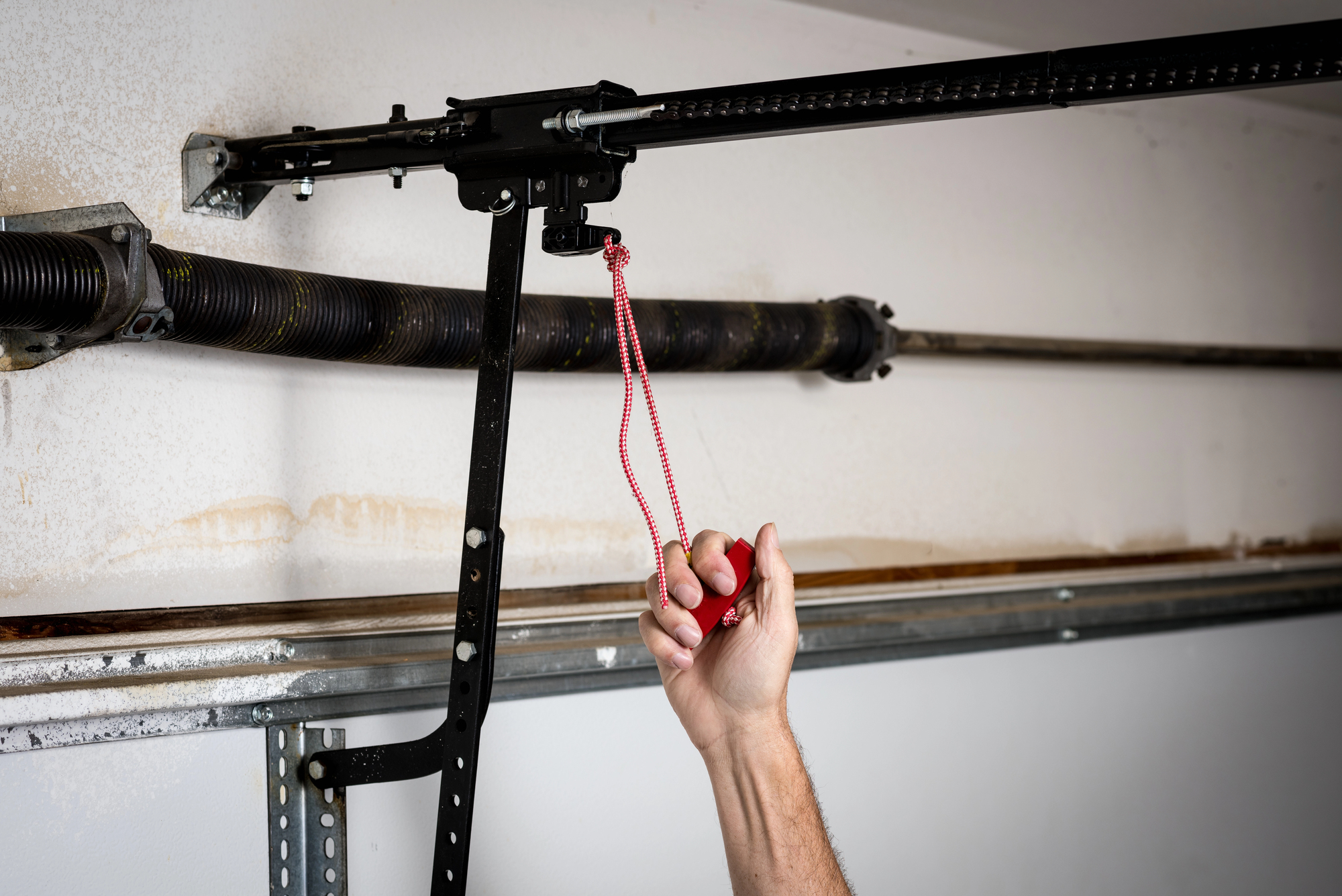 A hand pulls the red emergency release cord on a garage door opener mechanism mounted to the ceiling.