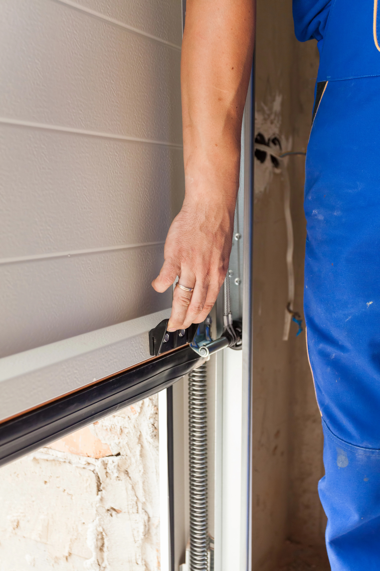 Person wearing blue work clothes adjusts or installs the bottom of a garage door, focusing on the metal hardware and spring mechanism.